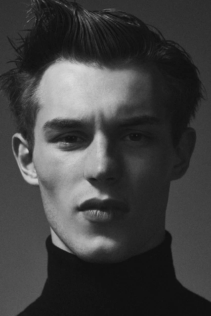 Monochrome headshot of a young adult male with dark hair styled upwards, wearing a black turtleneck sweater against a grey backdrop.