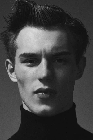 Monochrome headshot of a young adult male with dark hair styled upwards, wearing a black turtleneck sweater against a grey backdrop.