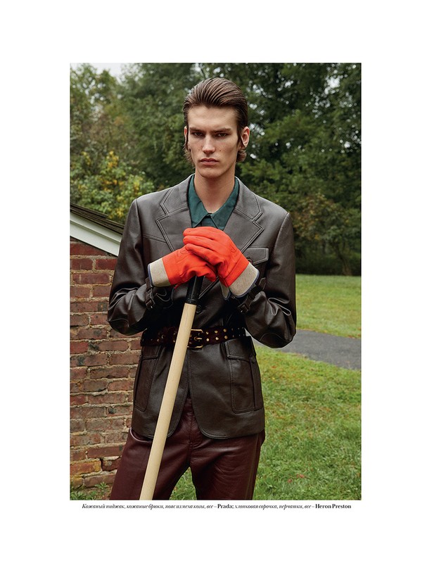Editorial fashion shot of a male model wearing a brown leather jacket and red gloves, posing with a shovel in a rural environment.