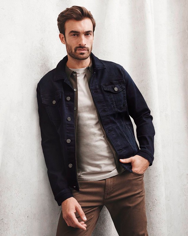 A man wearing a denim jacket and brown pants poses against a textured wall, capturing a blend of casual fashion and relaxed style.