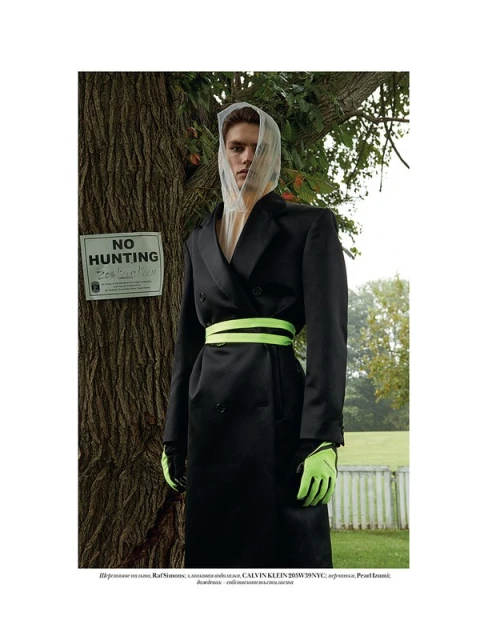 Fashion-forward model in a dark coat with neon green accents stands by a tree with a 'No Hunting' sign in an outdoor fashion shoot.