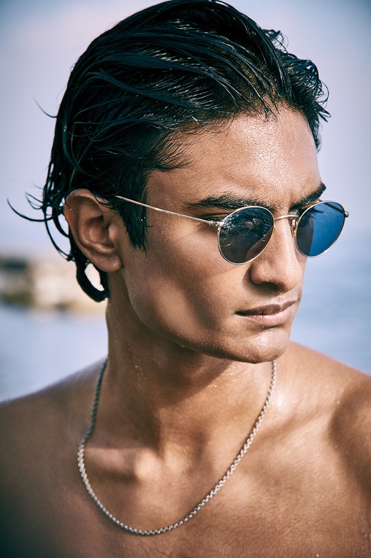A handsome adult man with dark, wet hair and sunglasses, wearing a chain necklace, poses outdoors against a blurred seascape background.