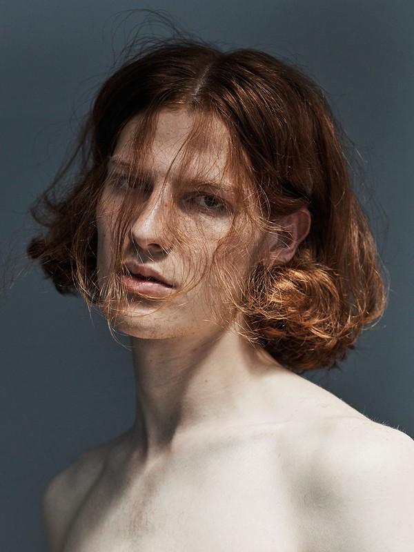 Portrait of a shirtless adult male with shoulder-length wavy auburn hair in a dimly lit photo studio against a solid grey backdrop.