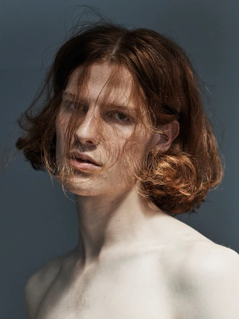 Portrait of a shirtless adult male with shoulder-length wavy auburn hair in a dimly lit photo studio against a solid grey backdrop.