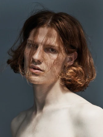 Portrait of a shirtless adult male with shoulder-length wavy auburn hair in a dimly lit photo studio against a solid grey backdrop.