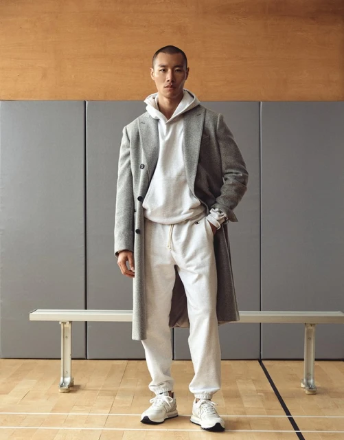 An Asian male model poses in a hoodie and overcoat in a gymnasium setting, creating a fashion-forward, athletic-inspired editorial shot.