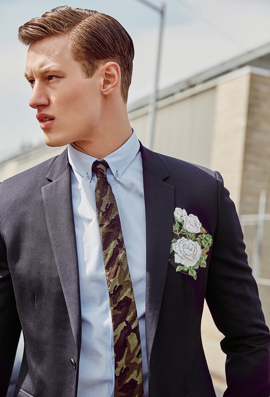 Stylish man in a dark suit, light blue shirt, and camo tie poses outdoors with neutral background. White floral pocket square.