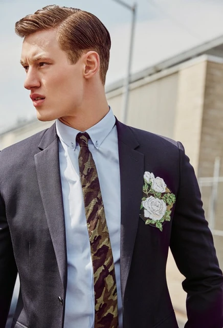 Stylish man in a dark suit, light blue shirt, and camo tie poses outdoors with neutral background. White floral pocket square.