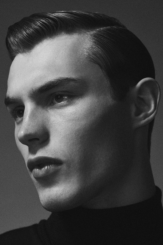 Black and white portrait of a young adult man with dark hair slicked back, wearing a dark turtleneck sweater against a plain backdrop.