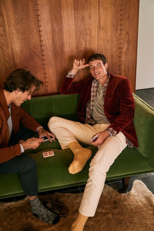 Two adult men relax on a green couch in a wood-paneled room. One man is seated while playing cards, the other is sitting and smiling.