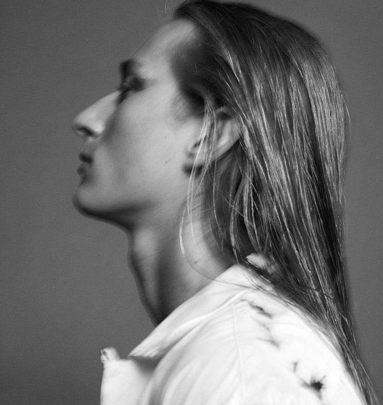 Profile portrait of a long-haired man with dark eye makeup wearing a white shirt against a neutral grey background.