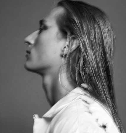 Profile portrait of a long-haired man with dark eye makeup wearing a white shirt against a neutral grey background.