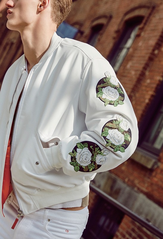 Close-up of a blond man wearing a white bomber jacket with floral designs and white jeans in an urban setting against a brick wall.