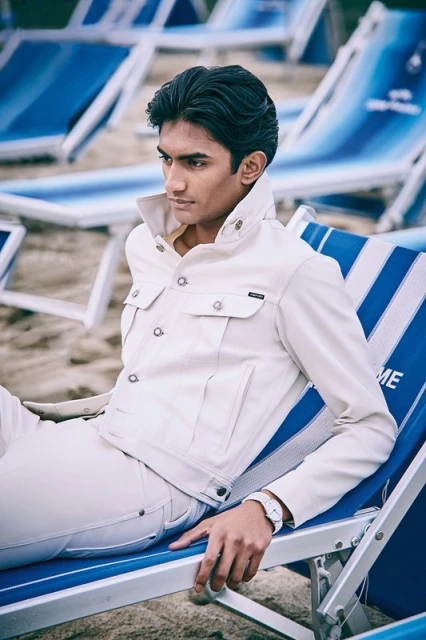 A young adult male in a cream-colored jacket sits on a blue-and-white striped lounge chair at the beach on a sunny day.