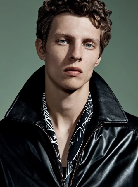 Close-up studio portrait of a young adult male with curly brown hair, wearing a black leather jacket and patterned shirt, against a solid green background.