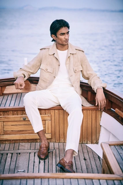 A young adult male sits on a wooden boat wearing a cream-colored jacket, white pants, and brown leather loafers, looking away toward the water.