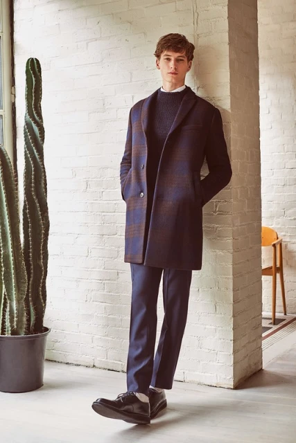 A young adult man in a blue and brown plaid overcoat and navy pants stands indoors against a white brick wall with a cactus.