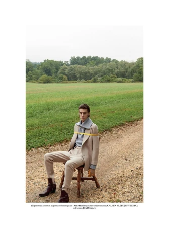 A young man seated on a wooden stool outdoors on a dirt path wearing a beige suit and boots with a grey sweater wrapped around his neck.