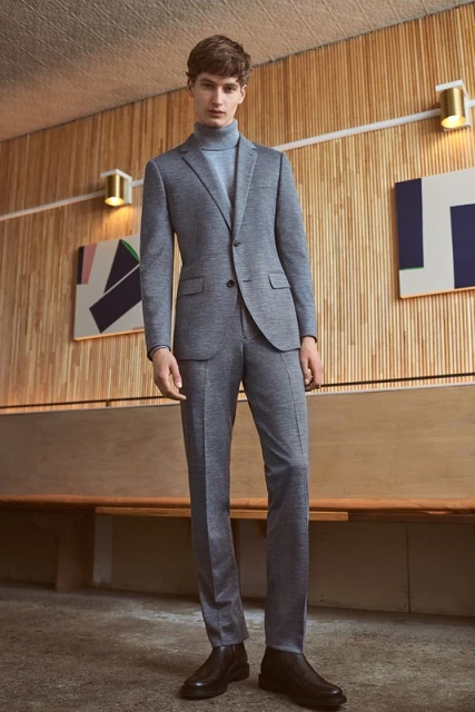 A full shot of a young man wearing a grey suit with a light blue turtleneck standing in front of a wooden wall with abstract paintings.