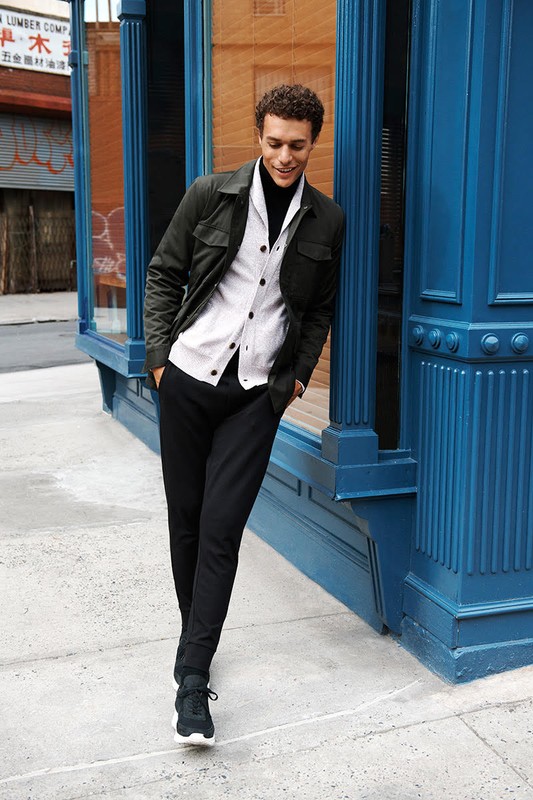 Man in jacket and button-down shirt leaning against a blue building on a sidewalk with trees in the background in an urban setting.