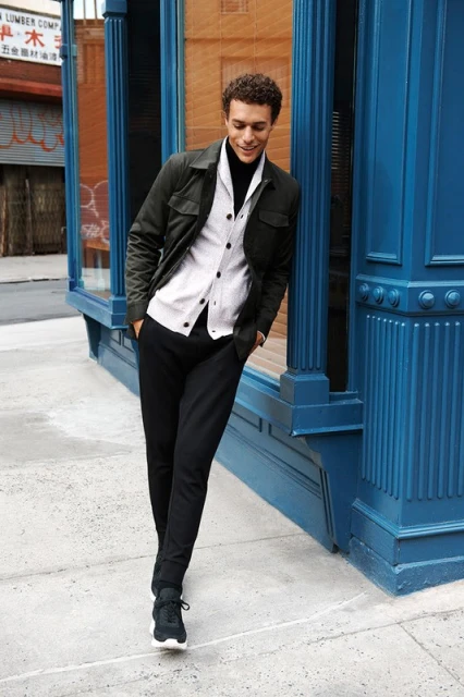 Man in jacket and button-down shirt leaning against a blue building on a sidewalk with trees in the background in an urban setting.