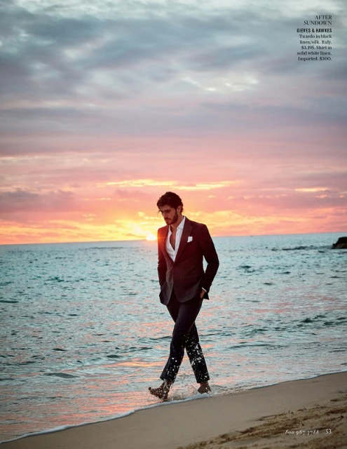 Man in a suit walks along the beach at sunset with ocean and cloudy sky visible in the background, wet sand, and barefoot.
