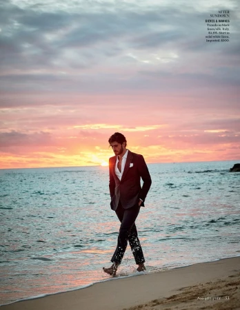 Man in a suit walks along the beach at sunset with ocean and cloudy sky visible in the background, wet sand, and barefoot.