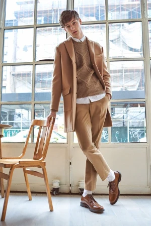 Male model in a camel coat and sweater standing with one hand on a wooden chair against large windows.