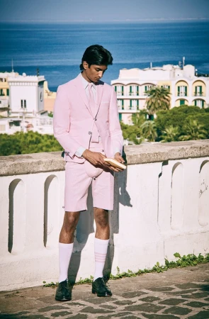 Man in pink suit and shorts poses in front of cityscape with the ocean. He's wearing white socks and dark shoes outdoors.