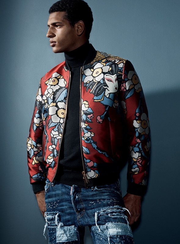 Fashion shot of a single adult male model wearing a red patterned bomber jacket with dark jeans, standing in a studio setting.