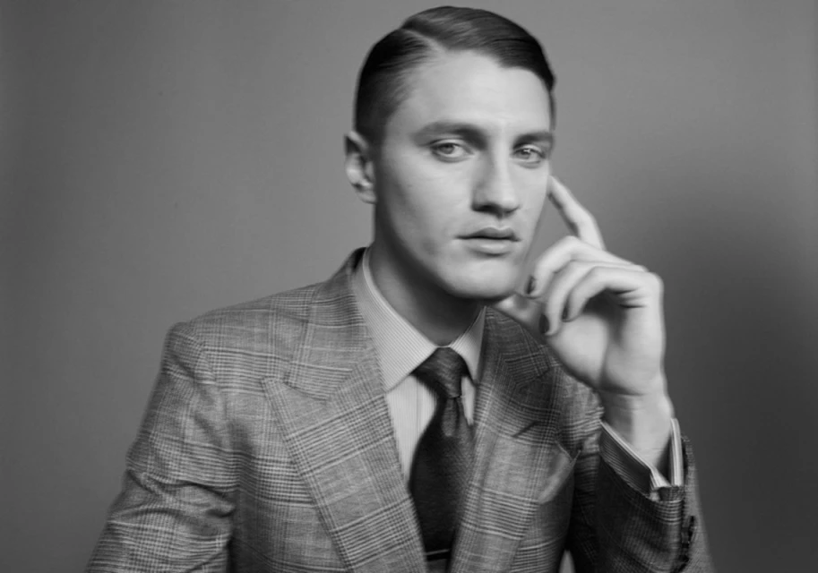Black and white portrait of an adult male in a suit and tie, with his hand near his face against a gray background.