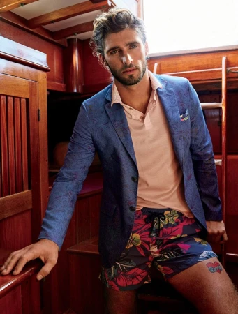 Stylish man in blazer and floral shorts on boat. Dark-haired male model poses in fashion shot, nautical setting, with wood background.
