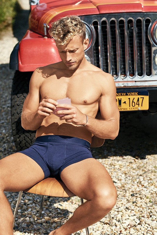 Shirtless blond male model in boxer briefs sits outside playing cards by a red Jeep, showcasing men's fashion and casual lifestyle.