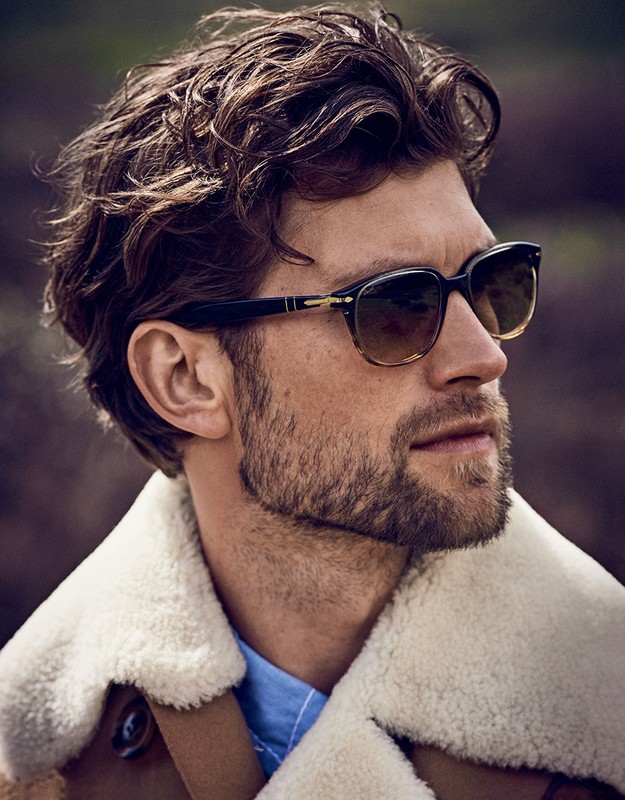 Portrait of an adult male wearing sunglasses and a jacket with a fur collar, gazing off-camera against an outdoor background.