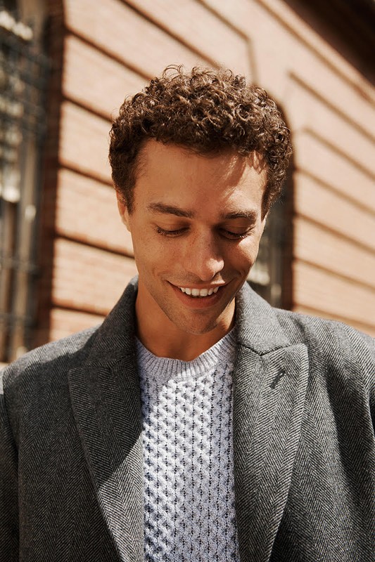 Smiling man with curly hair wearing a grey coat and textured light blue sweater stands outdoors against a brick wall.