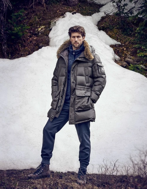 A man in a fur-trimmed parka and dark pants stands outdoors in a snowy, wooded environment, showcasing winter fashion apparel.