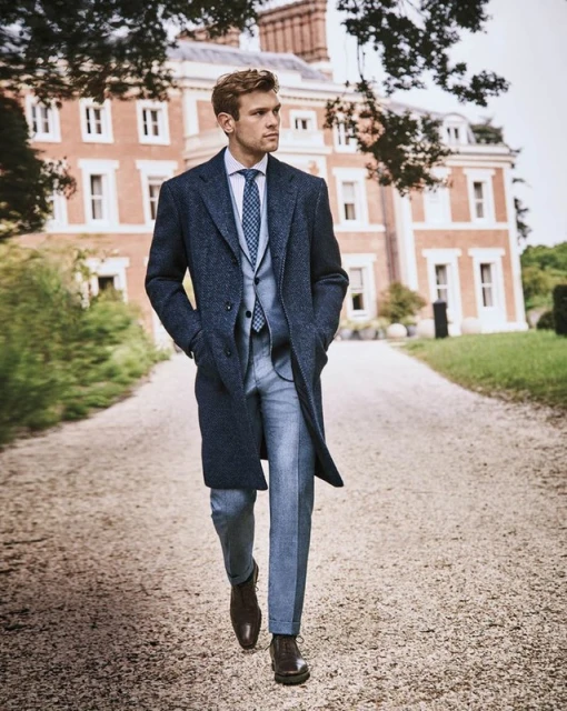A fair-skinned adult male in a blue suit and overcoat walks on a gravel path near a large brick building with trimmed green lawn.