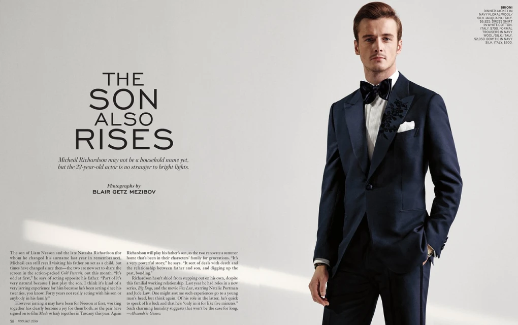Portrait of a young adult male in a navy blue floral dinner jacket with bow tie and white dress shirt against a plain background.