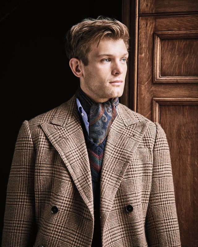 A blond male model poses indoors, wearing a plaid blazer, blue shirt, and patterned neck scarf, standing in front of a wood paneled door.