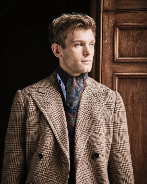 A blond male model poses indoors, wearing a plaid blazer, blue shirt, and patterned neck scarf, standing in front of a wood paneled door.