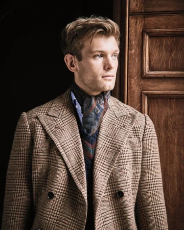 A blond male model poses indoors, wearing a plaid blazer, blue shirt, and patterned neck scarf, standing in front of a wood paneled door.