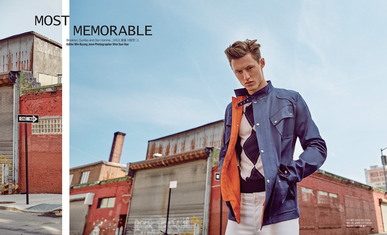 Male fashion model in blue jacket poses against urban backdrop with red brick buildings and blue sky, captured in a medium outdoor shot.