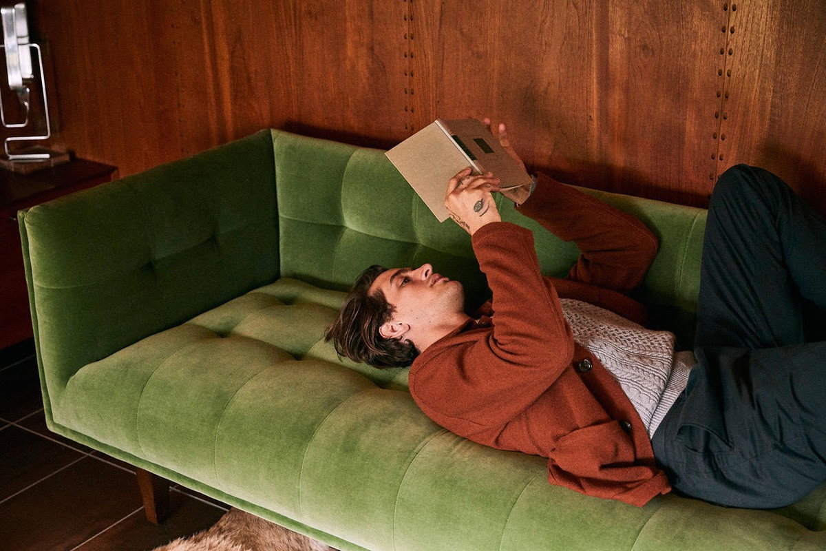 An adult man relaxes on a green velvet sofa, reading a book in a warmly lit indoor setting with wood paneled walls. He wears a brown jacket.