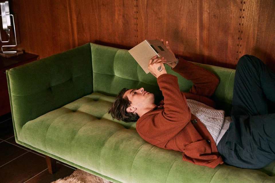 An adult man relaxes on a green velvet sofa, reading a book in a warmly lit indoor setting with wood paneled walls. He wears a brown jacket.
