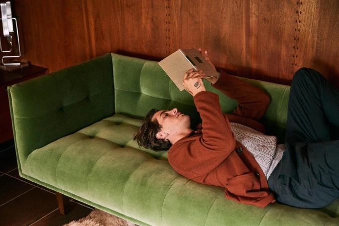 An adult man relaxes on a green velvet sofa, reading a book in a warmly lit indoor setting with wood paneled walls. He wears a brown jacket.