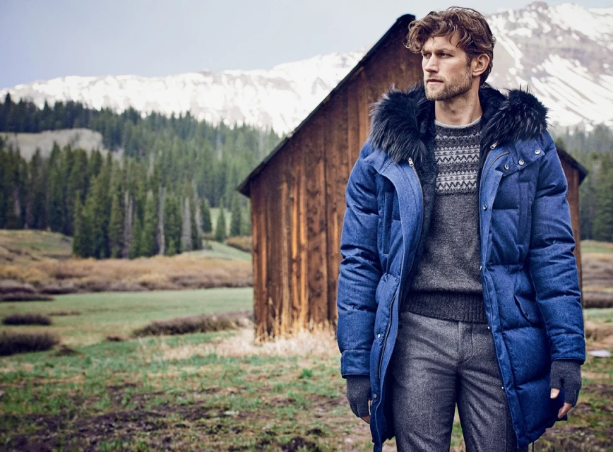 Outdoor portrait of a man in a blue puffer coat with fur trim, wearing a grey sweater and pants, standing in a mountain landscape.