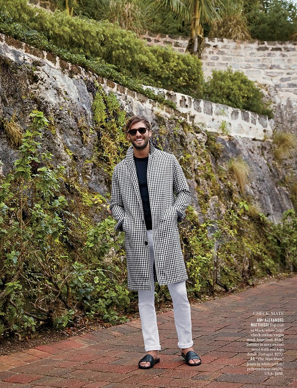 Smiling adult male with sunglasses posing outdoors on a brick path, wearing a black and white patterned coat and white pants.
