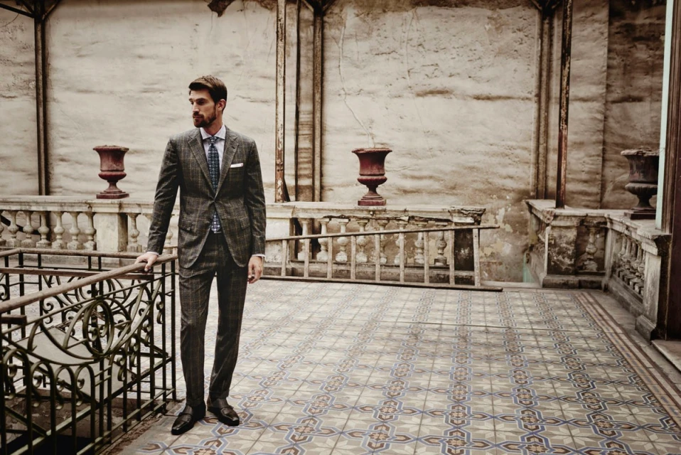 A handsome adult male models a plaid suit in an indoor environment with neutral walls and patterned floors, leaning on a decorative railing.