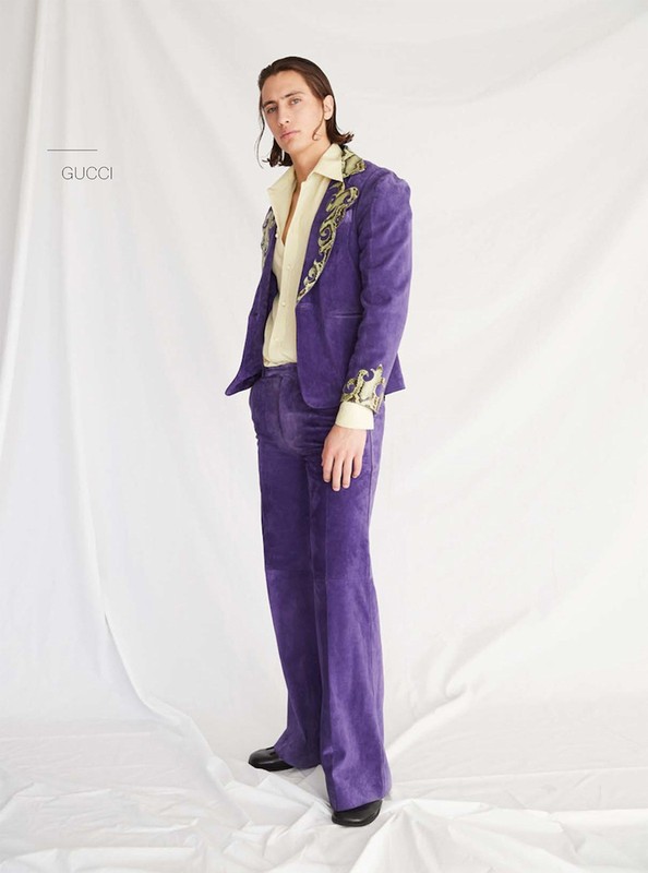 A young adult male posing in a purple velvet suit with floral embroidery, worn with a collared shirt, stands indoors against a white backdrop.