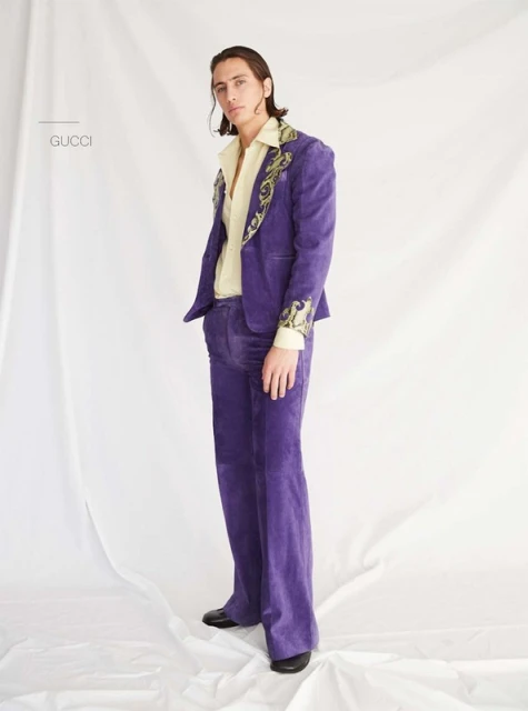 A young adult male posing in a purple velvet suit with floral embroidery, worn with a collared shirt, stands indoors against a white backdrop.